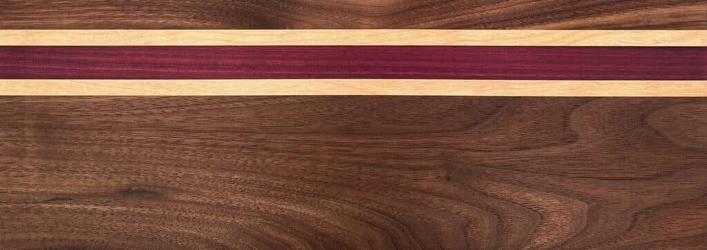A walnut cutting board, with maple and purpleheart stripes running horizontally. The maple strips are half the width of the purpleheart and flank it on both sides. This is at the top third of the cutting board